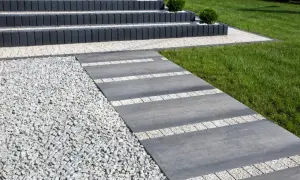 Nowoczesny chodnik przed domem z szarej płyty i białego gresu, otoczony żwirem i zielonym trawnikiem.