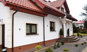 Nowy dom z białymi ścianami i brązowym dachem. Wokół domu widać zadbany ogród z kamiennym podjazdem i ozdobnymi krzewami. Cokół wokół domu jest w kolorze ceglastym.