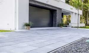 Nowoczesny, minimalistyczny pomysł na wjazd do posesji z szarej kostki brukowej i kamyczków. Elegancka brama garażowa i biała elewacja.
