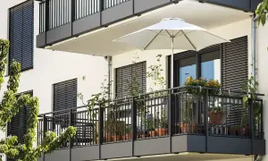 Nowoczesny balkon z białym parasolem i licznymi roślinami w doniczkach. Ciemna balustrada i okna z żaluzjami tworzą elegancką całość.