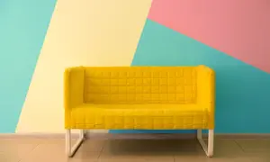 Żółta kanapa na tle geometrycznej ściany w kolorach: miętowym, żółtym i różowym. Świetne inspiracje jak pomalować pokój!