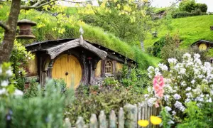 Uroczy dom hobbita z żółtymi drzwiami, porośnięty trawą, otoczony kwitnącymi kwiatami i zielenią.