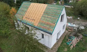 Budowa domu w stylu wiejskim. Dach w trakcie prac, widoczne drewniane łaty i folia dachowa. Wokół materiały budowlane i zieleń.