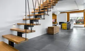 Nowoczesna klatka schodowa w domu piętrowym, z drewnianymi stopniami i czarnymi linami. Obok salon z kominkiem i szarą kanapą.
