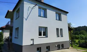 Jasnoszary dom z lat 80. z balkonem i ciemnoszarą podstawą, otoczony zielenią.