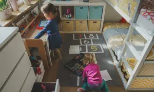 Dwie dziewczynki bawią się w przytulnym pokoju dla dwójki nastolatków. Jedna rysuje przy biurku, druga siedzi na dywanie z kredkami.