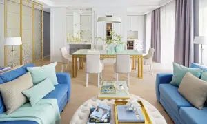 Eleganckie styl hampton aranżacje: błękitne sofy, złoty stół jadalniany, jasne krzesła i dekoracje tworzą przytulną przestrzeń.
