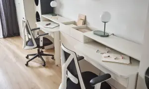 Mały pokój dla 3 dzieci z dwoma biurkami i krzesłami. Na biurkach lampki, zeszyty i kredki.