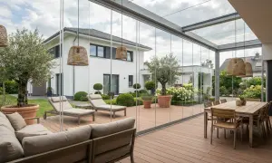 Letnie dekoracje tarasu: wygodna sofa, leżaki i stół jadalniany pod szklanym zadaszeniem, z widokiem na ogród.