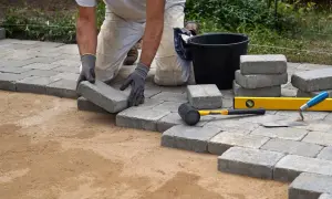 Ręce w rękawiczkach układają kostkę brukową, tworząc ścieżkę. Narzędzia ogrodnicze i stosy kostki sugerują inspiracje do własnego projektu.