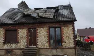 Zniszczony dach domu z kamienia wapiennego. Obok stoi wóz strażacki, a przy nim strażacy.