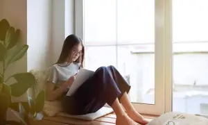 Dziewczynka w okularach siedzi na wygodnym siedzisku pod oknem, rysując coś w szkicowniku.
