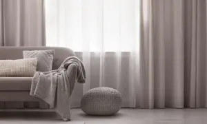 Miękka sofa z poduszkami i pledem, obok okrągła pufa. Elegancka dekoracja okien z zasłonami tworzy przytulną atmosferę.