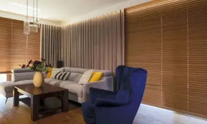 Elegancki salon z szarym narożnikiem, granatowym fotelem i drewnianymi żaluzjami oraz zasłonami w ciepłych odcieniach.
