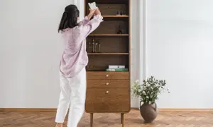 Kobieta układa książki na półce w stylu mid century modern. Drewniany mebel z zaokrąglonymi krawędziami i mosiężnymi uchwytami.