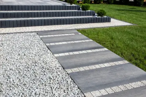 Nowoczesny chodnik przed domem z szarej płyty i białego gresu, otoczony żwirem i zielonym trawnikiem.