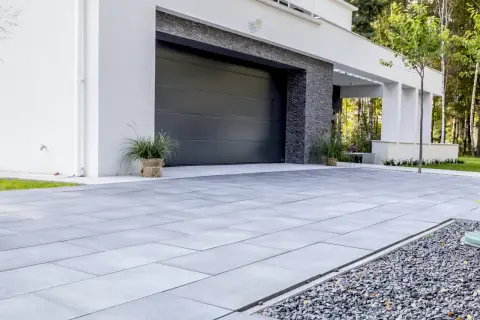Nowoczesny, minimalistyczny pomysł na wjazd do posesji z szarej kostki brukowej i kamyczków. Elegancka brama garażowa i biała elewacja.