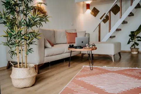 Przytulne, **małe mieszkanie w stylu boho** z beżową kanapą, drewnianym stolikiem i roślinami. Schody prowadzą na piętro.