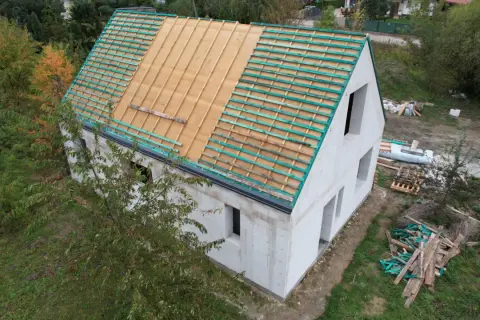 Budowa domu w stylu wiejskim. Dach w trakcie prac, widoczne drewniane łaty i folia dachowa. Wokół materiały budowlane i zieleń.