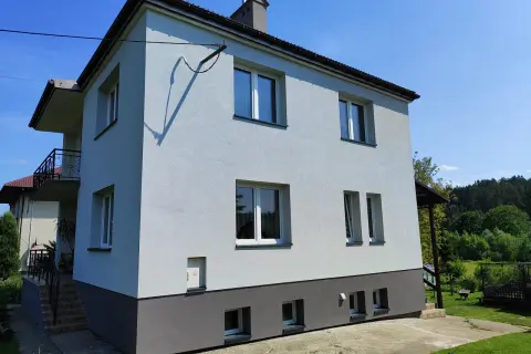 Jasnoszary dom z lat 80. z balkonem i ciemnoszarą podstawą, otoczony zielenią.