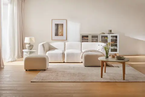 Jasny, przytulny salon z beżową kanapą i pufą. Na podłodze dywan i drewniana podłoga.