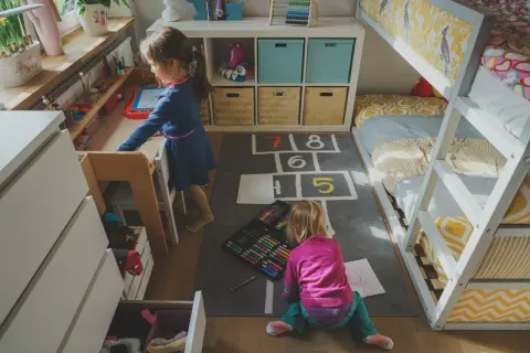 Dwie dziewczynki bawią się w przytulnym pokoju dla dwójki nastolatków. Jedna rysuje przy biurku, druga siedzi na dywanie z kredkami.
