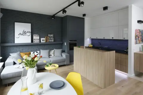 Nowoczesny salon z aneksem kuchennym w bloku, urządzony w stylu loftowym. Szara cegła, drewno i jasne meble tworzą przytulną przestrzeń.
