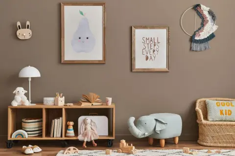 Przytulny pokój zabaw dla dzieci w domu. Na ścianie wiszą obrazki z gruszką i napisem "Small steps every day". Na półce stoi lampka, pluszowa owieczka i książki. Obok siedzi lalka-króliczek. Na podłodze leżą drewniane klocki i puf w kształcie słonia.