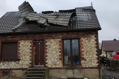 Zniszczony dach domu z kamienia wapiennego. Obok stoi wóz strażacki, a przy nim strażacy.