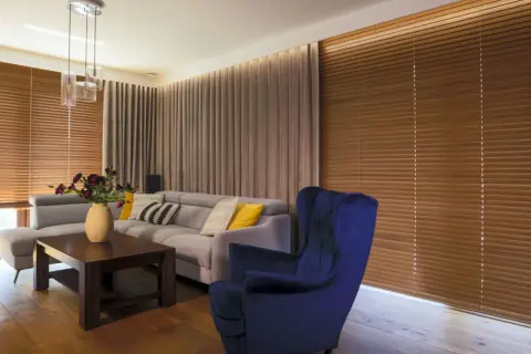 Elegancki salon z szarym narożnikiem, granatowym fotelem i drewnianymi żaluzjami oraz zasłonami w ciepłych odcieniach.