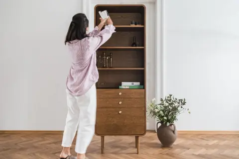 Kobieta układa książki na półce w stylu mid century modern. Drewniany mebel z zaokrąglonymi krawędziami i mosiężnymi uchwytami.