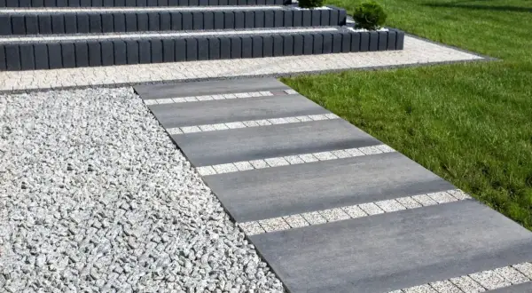 Nowoczesny chodnik przed domem z szarej płyty i białego gresu, otoczony żwirem i zielonym trawnikiem.