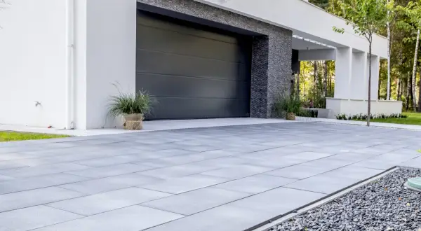 Nowoczesny, minimalistyczny pomysł na wjazd do posesji z szarej kostki brukowej i kamyczków. Elegancka brama garażowa i biała elewacja.