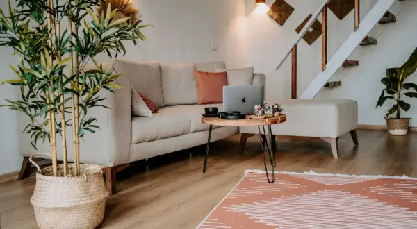 Przytulne, **małe mieszkanie w stylu boho** z beżową kanapą, drewnianym stolikiem i roślinami. Schody prowadzą na piętro.