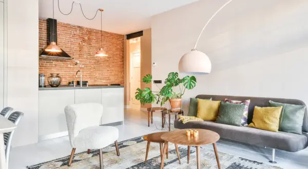 Nowoczesne inspiracje mieszkaniowe: salon z szarą kanapą, biały fotel, stoliki kawowe, rośliny i kuchnia z ceglaną ścianą.