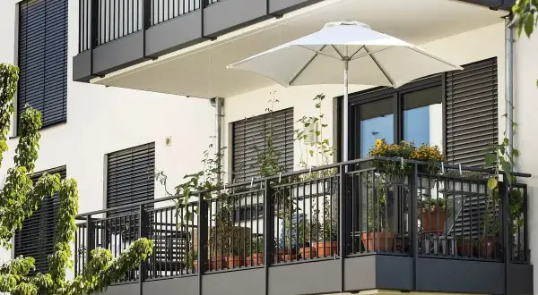 Nowoczesny balkon z białym parasolem i licznymi roślinami w doniczkach. Ciemna balustrada i okna z żaluzjami tworzą elegancką całość.