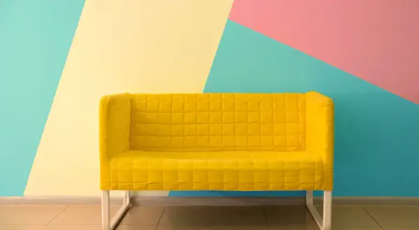 Żółta kanapa na tle geometrycznej ściany w kolorach: miętowym, żółtym i różowym. Świetne inspiracje jak pomalować pokój!