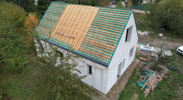 Budowa domu w stylu wiejskim. Dach w trakcie prac, widoczne drewniane łaty i folia dachowa. Wokół materiały budowlane i zieleń.