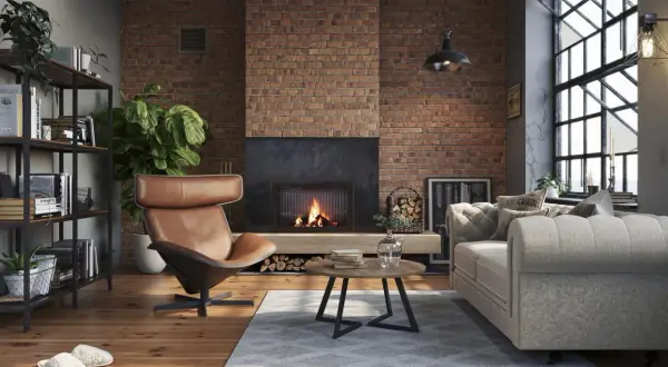Przytulny, **soft loft** z palącym się kominkiem, skórzanym fotelem i szarą kanapą. Ceglana ściana i drewniana podłoga tworzą ciepłą atmosferę.