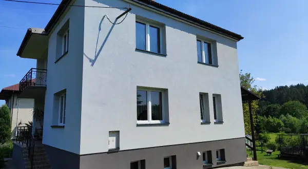 Jasnoszary dom z lat 80. z balkonem i ciemnoszarą podstawą, otoczony zielenią.