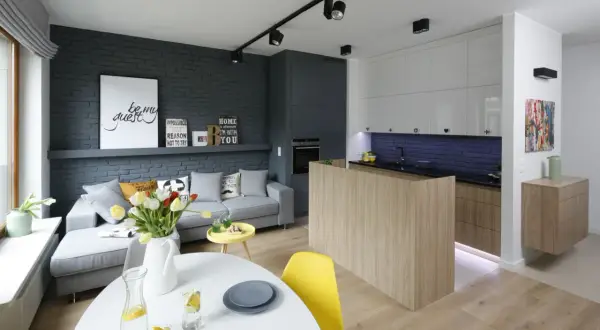 Nowoczesny salon z aneksem kuchennym w bloku, urządzony w stylu loftowym. Szara cegła, drewno i jasne meble tworzą przytulną przestrzeń.