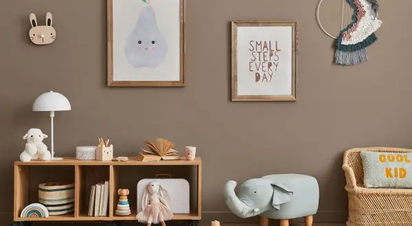 Przytulny pokój zabaw dla dzieci w domu. Na ścianie wiszą obrazki z gruszką i napisem "Small steps every day". Na półce stoi lampka, pluszowa owieczka i książki. Obok siedzi lalka-króliczek. Na podłodze leżą drewniane klocki i puf w kształcie słonia.