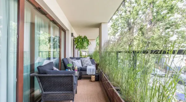 Przytulny balkon w bloku z wygodną sofą i stolikiem, otoczony bujną zielenią traw ozdobnych. Idealne miejsce na relaks.