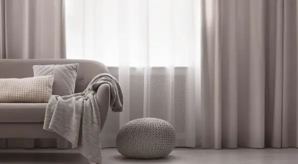 Miękka sofa z poduszkami i pledem, obok okrągła pufa. Elegancka dekoracja okien z zasłonami tworzy przytulną atmosferę.