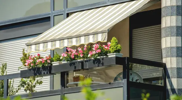 Pasiasty markiza nad balkonem w bloku, z kwitnącymi różowymi kwiatami w skrzynkach, to świetny sposób, jak osłonić balkon.