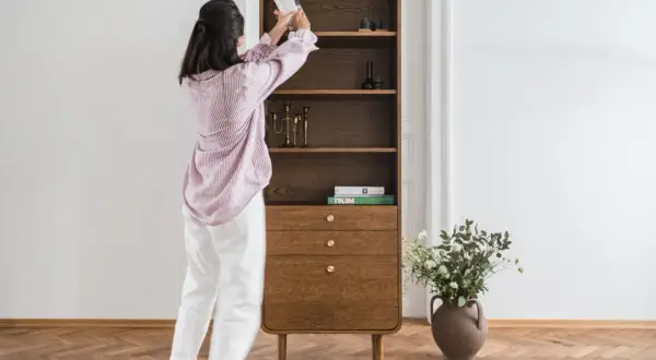 Kobieta układa książki na półce w stylu mid century modern. Drewniany mebel z zaokrąglonymi krawędziami i mosiężnymi uchwytami.