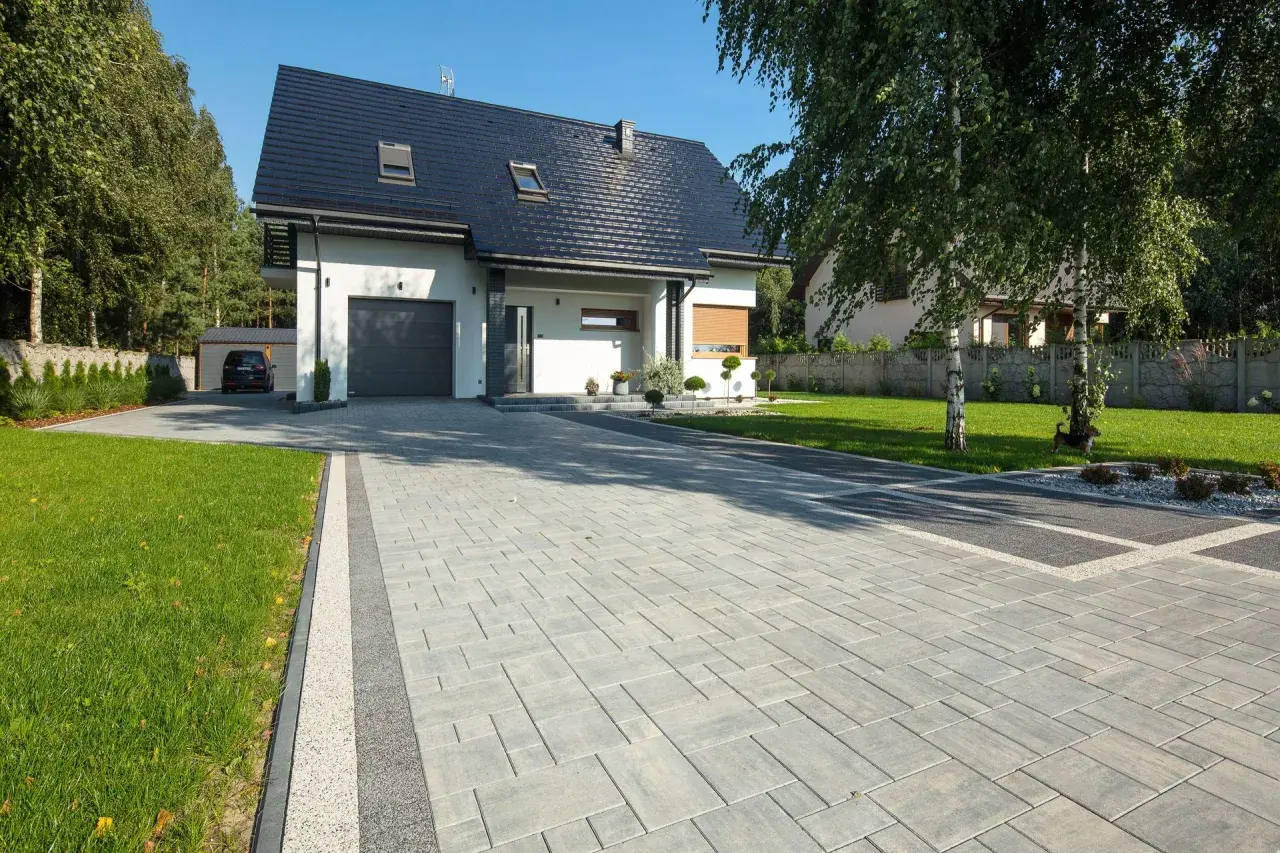 Nowy podjazd do domu z kostki brukowej, prowadzący do garażu. W tle nowoczesny dom i zadbany ogród.
