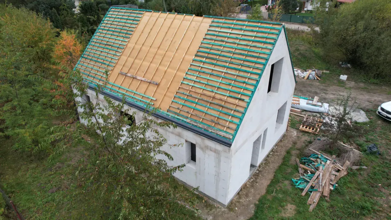 Budowa domu w stylu wiejskim. Dach w trakcie prac, widoczne drewniane łaty i folia dachowa. Wokół materiały budowlane i zieleń.