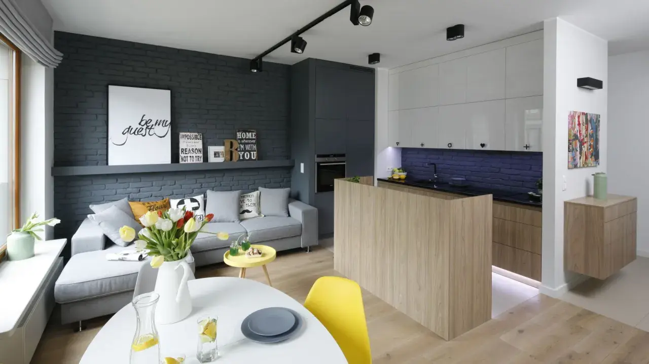 Nowoczesny salon z aneksem kuchennym w bloku, urządzony w stylu loftowym. Szara cegła, drewno i jasne meble tworzą przytulną przestrzeń.
