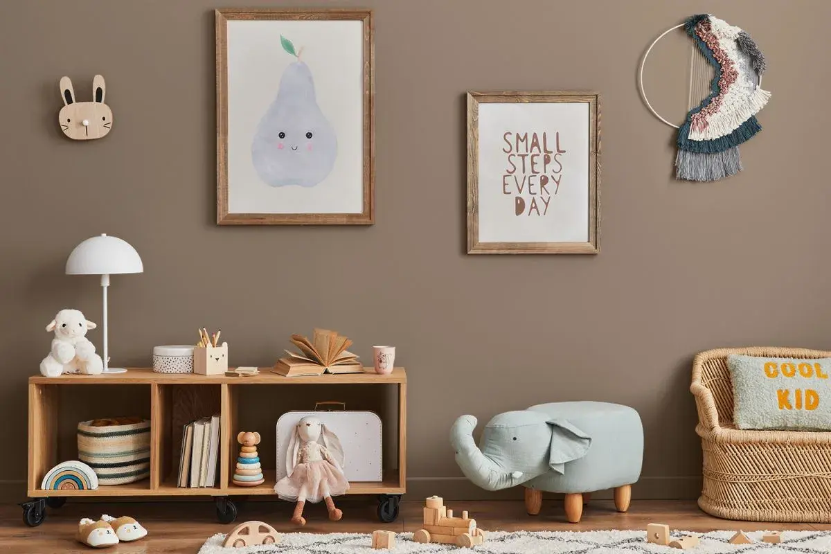 Przytulny pokój zabaw dla dzieci w domu. Na ścianie wiszą obrazki z gruszką i napisem "Small steps every day". Na półce stoi lampka, pluszowa owieczka i książki. Obok siedzi lalka-króliczek. Na podłodze leżą drewniane klocki i puf w kształcie słonia.